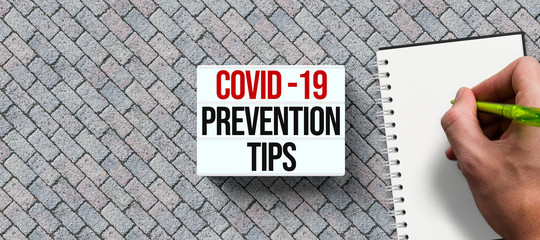 lightbox with text COVID-19 PREVENTION TIPS and hand with pen over a notepad on stone background