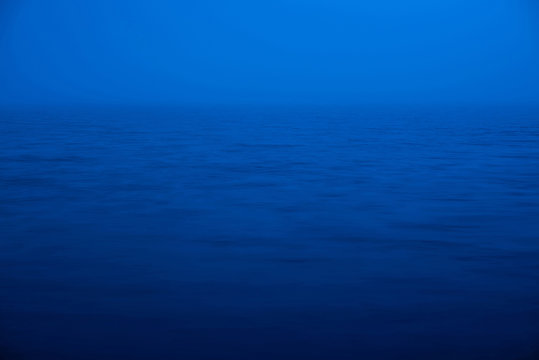 Water ripple nature background. Natural texture of sea of blue classic color. Deep blue calm water in dusk close-up. Meditative image of dense fog above lake. Soft light glitters on water surface.