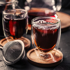 Cup of freshly brewed black tea, dark moody, tea ceremony. Hot water is poured from the kettle into a flass cup on wooden tray, black concrete background.