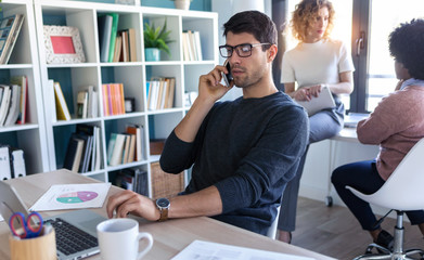 Handsome young entrepreneur talking on mobile phone while working with laptop in the office.