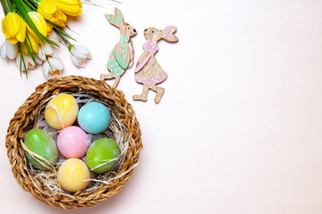 Easter card template. Top view on colorful eggs with straw in a wicker basket, a bouquet of spring flowers and a couple of wooden easter bunnies flat lay on a pink background. Large space for text.