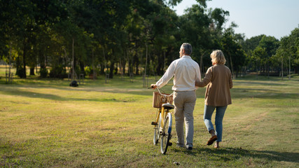 The behind of Caucasian elderly couples walking with a bicycle in the natural autumn sunlight garden feel cherish and love, concept elderly love, warm family, happy retirement, retirement lifestyle.