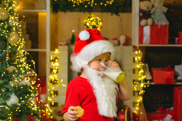 Fototapeta na wymiar Portrait of Santa Claus Drinking milk from glass and holding cookies. Little Santa Claus kid with beard and mustache. Santa boy in Santa hat. Christmas for kid.