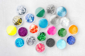 Creative flat lay overhead composition with sequins, glitter, mica and shimmer for nail design in plastic jars on a light wooden background. Top view.