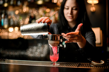 Fototapeta na wymiar Woman bartender pours cocktail from shaker into glass using sieve.