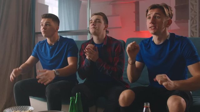 A group of men in blue t-shirts. Watch a football match with friends sitting on the couch and cheer and applaud while looking directly at the camera