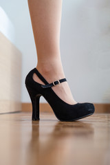 Caucasian female legs close up shot wearing black high heel shoes inside on wooden tile pavement.