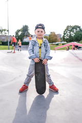 little boy 4-5 years old, stands with a skateboard, in summer in city on sports field sits. Casual wear denim and baseball cap. Emotions of positive and joy smile.