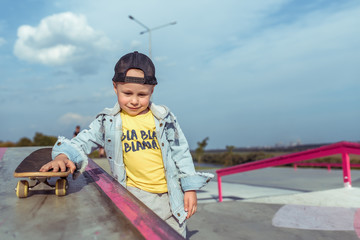 little boy 4-5 years old, rolls a skateboard, in summer in city on sports field sits. Casual wear denim and baseball cap. Free space for copy text. Emotions of positive and joy smile.
