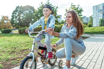 Young family, mother woman, little boy 3-5 years old son, summer in city. Learning to ride bicycle, balance training, help support of parent in raising child. Emotions of positive and happiness joy.
