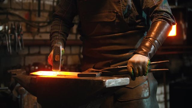 Blacksmith working in the workshop - man heating up the longer piece of metal in the furnace and hitting it with a hammer