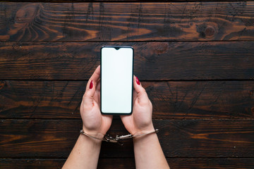 Top view woman hands in handcuffs hold a smartphone on a wooden background. The concept of internet and gadget dependency.