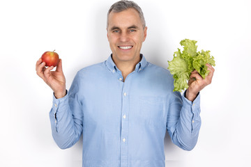 adult man with grey hair wearing blue shirt standing with apple in one hand and salad in another on...