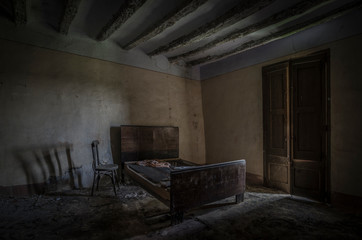 Fototapeta na wymiar Dormitorio de casa abandonada con cama y silla en la penumbra iluminada por una ventana.
