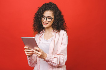 Beautiful curly young girl smiling using and looking at tablet isolated over red background. Copy...