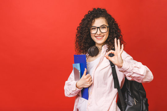 Young Curly Student Woman Wearing Backpack Glasses Holding Books And Tablet Over Isolated Red Background.  Ok Sign.