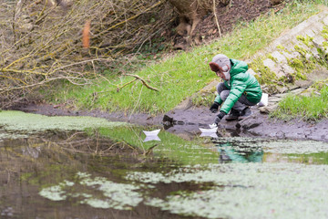 Obraz premium A boy in an autumn jacket and hat stands on the riverbank and looks at paper boats.