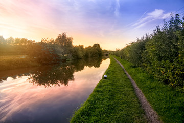 Looking along the tow path of the Grand Union Canal in Northamptonshire with a colorful sunset behind the trees on the opposite bank with the orange and red sky reflecting in the still water