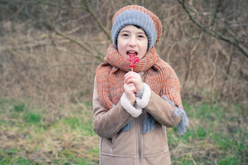 Portrait of a beautiful boy in a knitted hat with a scarf on the background of autumn (winter) nature. In the hands of the boy holds a sweet candy in the shape of rooster.
