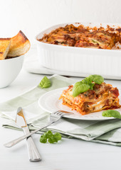 A close up of a slice of lasagna with the pan of lasagna in behind and a dish of garlic toast.
