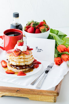 A tray table of breakfast items including pancakes, coffee and strawberries with a card and flowers for Mother's Day.