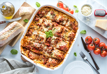 A top down view of a freshly baked lasagna cut into slices ready for serving and surrounded by Italian ingredients.