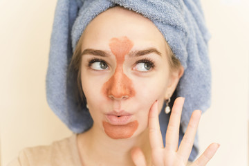 A beautiful girl with a towel on her head makes a multi mask on the t-zone of orange color with a scrub. The cute blonde is having fun doing a home care, moisturizing and smoothing facial mask
