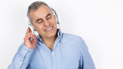 adult handsome man with grey hair wearing blue shirt listens to music in headphones standing on isolated white background, positive vibes concept