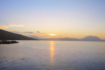 Obraz premium Serene bay on Corfu coast with the sun setting behind distant mountains