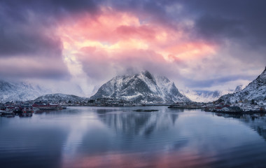 Aerial view of snowy mountain, village on sea coast, blue sky with red clouds at sunset in winter. Top view of Reine, Lofoten islands, Norway. Landscape with rocks, houses, rorbu, reflection in water