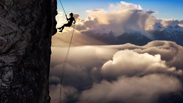 Cinemagraph Loop. Man (Unrecognizable) Rappelling from Cliff. Beautiful aerial view of the clouds and mountains during a colorful and vibrant sunset or sunrise. Landscape from BC, Canada.