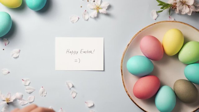hand puts a greeting card on the easter decorated table. colorfull eggs on plate and blossom branch on blue background. holiday concept. top view, 4K.