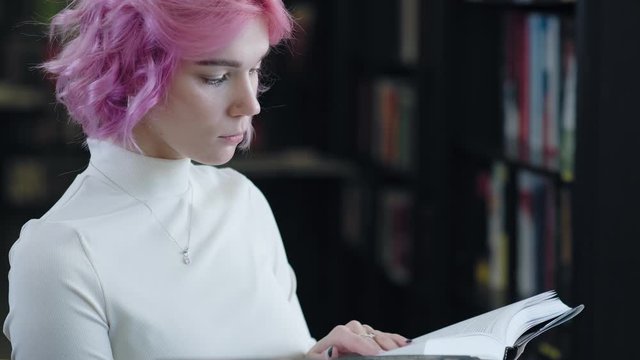 Modern woman with pink hair is reading a book in the bookshop.