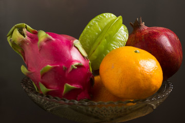 Fototapeta na wymiar A lot of tropical fruits in a glass vase. Exotic fruits: averrhoa carambola, Pitahaya, Mango, tangerines, pomegranate fruit. Gray background