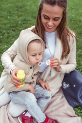 Happy woman mom, little boy plays inflates soap bubbles, casual warm clothes hood. Autumn day summer on grass. Weekend breaks, caring parenting childcare. Emotions Happiness joy fun smile.
