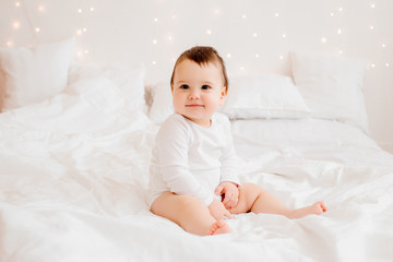 healthy baby boy 10 months old in white clothes smiling sitting on white bed linen in bed, space for text