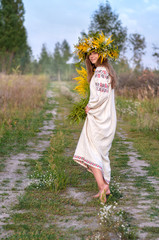 Young beautiful woman in a wreath of wildflowers and a ancient national clothes walking barefoot on the path. Ukrainian culture, holiday of Ivana Kupala