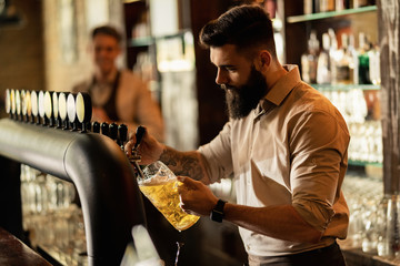 Young bartender pouring draft beer form beer tap in a pub.