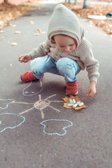 Fototapeta premium little boy in beige jumper with hood, autumn day in summer in city, draws sun on asphalt with colorful chalks. Creative concept idea and creativity.
