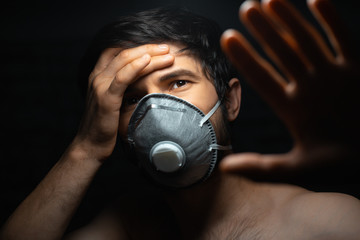 Virus concept. Dramatic dark portrait of young man wearing protective flu mask, holding head with one hand and showing stop gesture with another one. Background of black color.