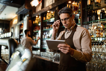 Mid adult bartender using touchpad while working in a pub. - obrazy, fototapety, plakaty