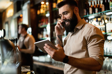Young happy bartender using touchpad while talking on the phone.