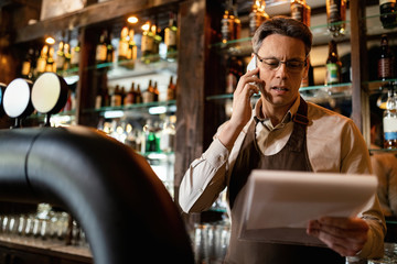 Mid adult barista making phone call while going through inventory list in a bar.