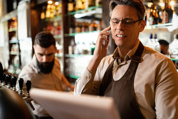 Smiling barista making a phone call while reading inventory list in a bar.