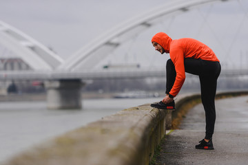 Young sporty man in orange windbreaker preparing for jogging on a cloudy day tying his shoes