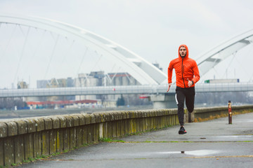 Handsome young athlete man running fast along river in orange windbreaker jacket. Extreame weather sport. Running on a rainy day.