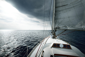 White sloop rigged yacht sailing near the coast of Maine, Southwest Harbor, USA. A view from the deck to the bow, mast and sails. Dark blue storm sky, sun rays through the clouds