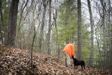 A man walking in the woods stops to pet his black Labrador dog. The man is wearing an orange jacket. There is room for your text on the left side of the frame.