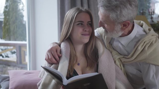 Senior Caucasian man in love talking with young pretty Caucasian woman reading book. Happy loving couple with age difference enjoying time together indoors. Lifestyle, love, romance, tranquility.
