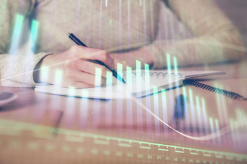 Hands writing information about stock market in notepad. Forex chart holograms in front. Concept of research. Multi exposure